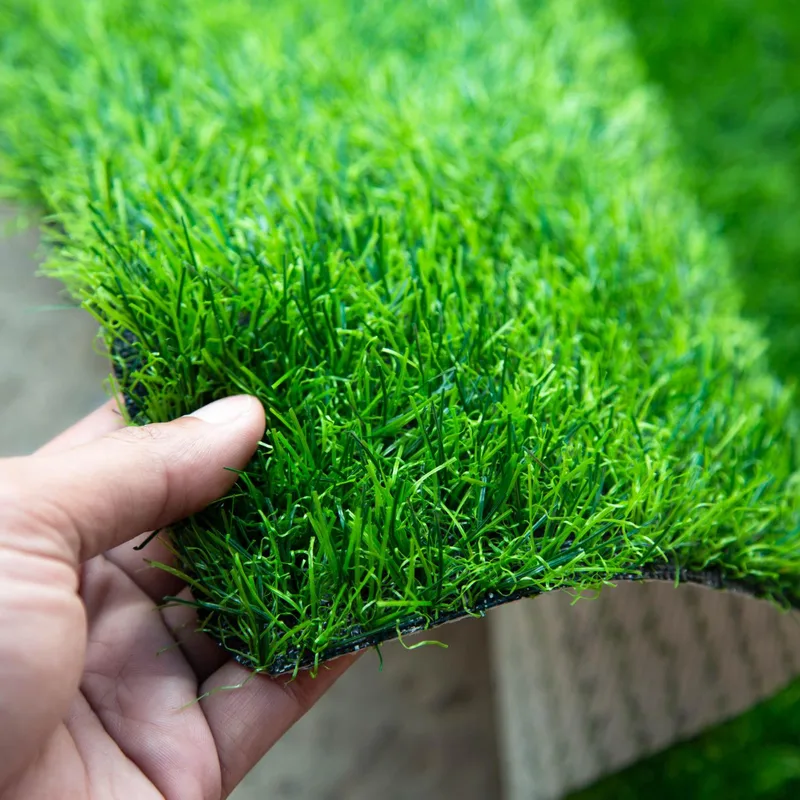 Hand holding up a sample of dense green artificial grass with a red track surface beside it