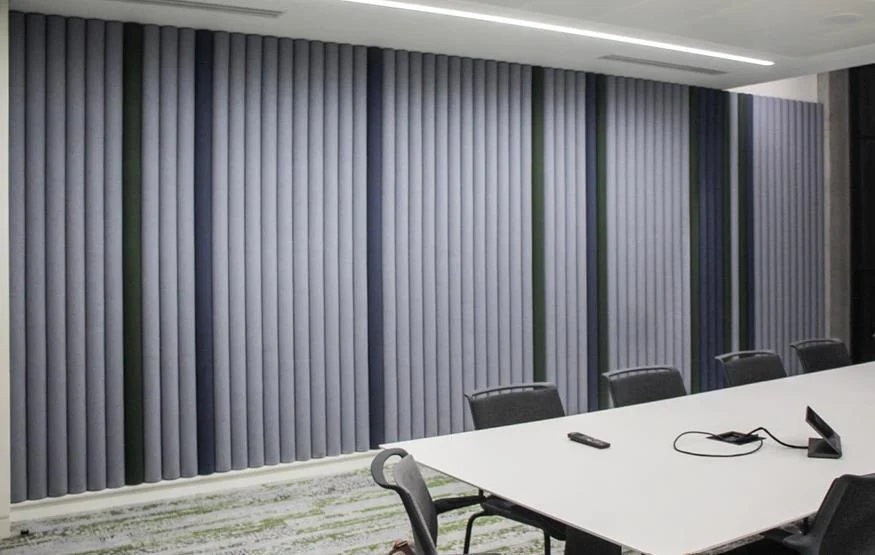 Conference hall with a long white table and black chairs in front of a wall of tall, dark vertical acoustic panels under linear lights