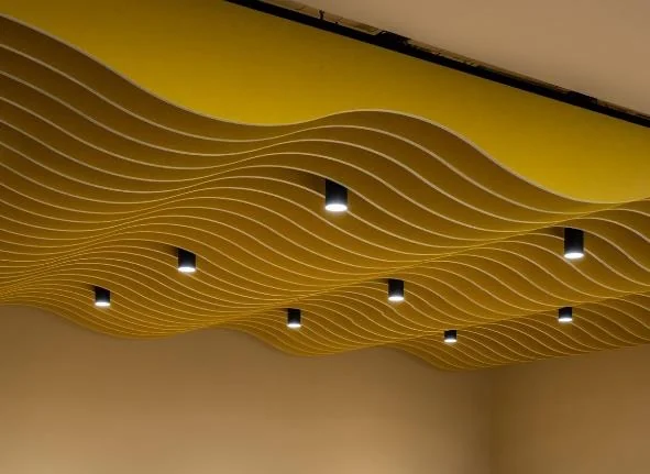 Ceiling of flowing yellow wave-shaped acoustic panels with round black downlights set between the waves