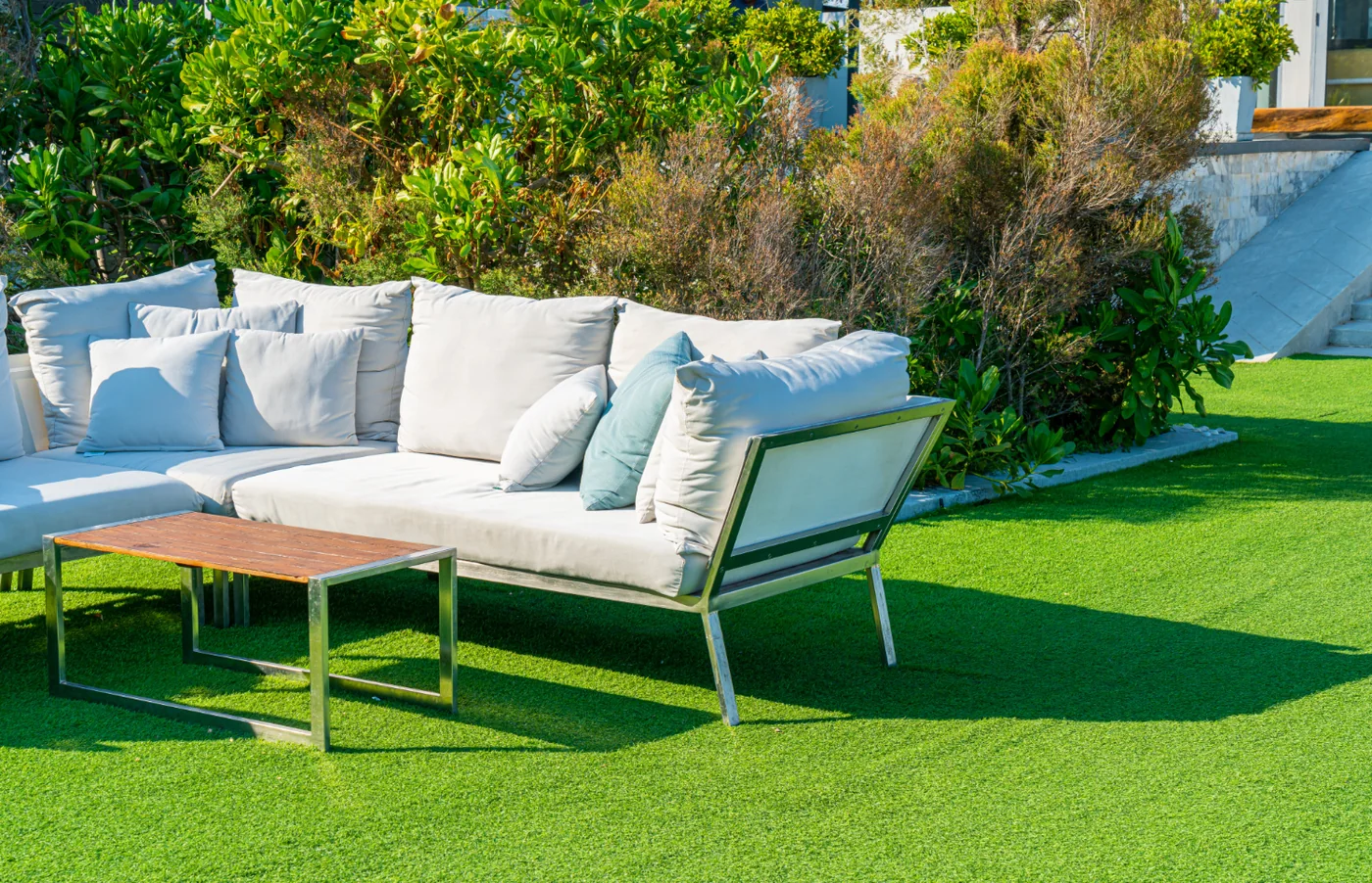 White outdoor sofa with white and teal cushions and a metal-framed timber-top table on a green artificial lawn, with shrubs behind
