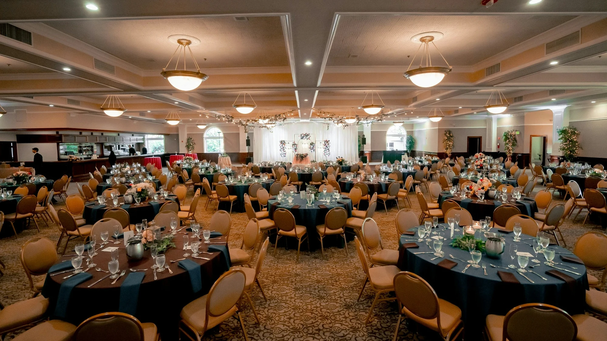 Large banquet hall with round tables set with navy cloths and gold chairs on a patterned carpet under a coffered ceiling with chandeliers