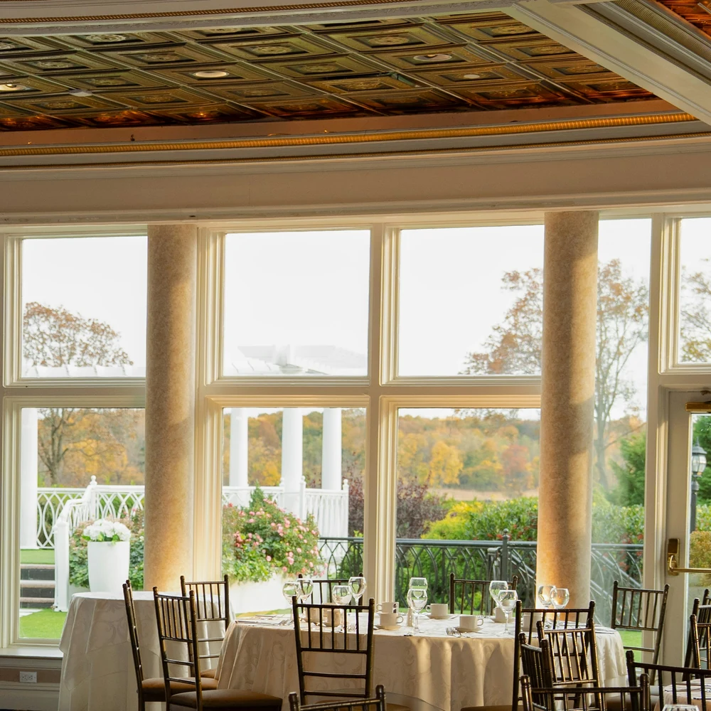 Sunlit banquet room with round tables, cane-back chairs and tall windows looking onto a garden