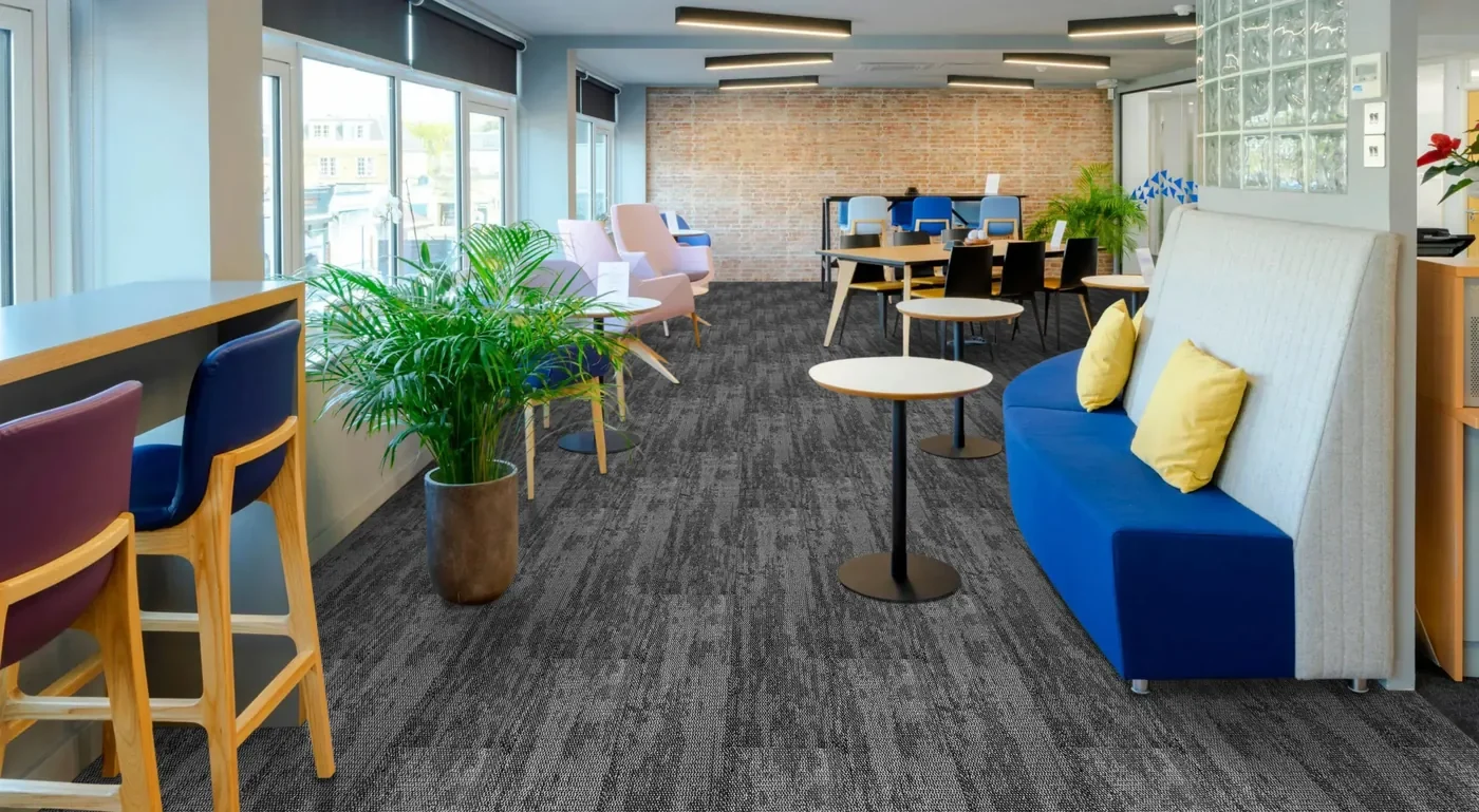 Open-plan office breakout area with dark grey carpet planks, a blue sofa, bar stools and potted plants