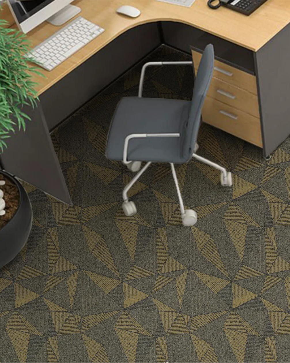 Office desk corner with a grey chair on a dark carpet tile floor patterned with gold triangles, and a plant beside the desk