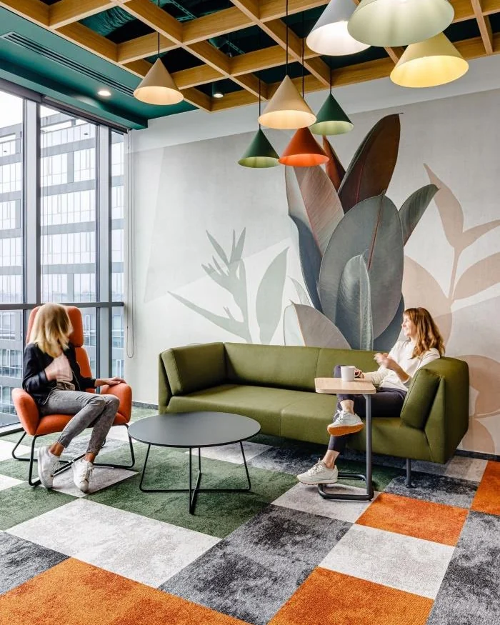 Office lounge with a green sofa and two people talking, on carpet tiles in blocks of orange, grey and green beneath pendant lights