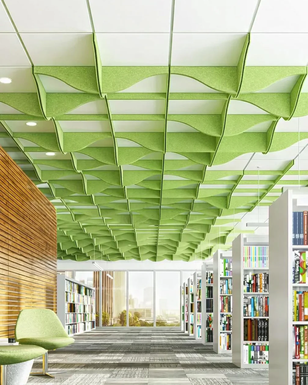 Library aisle between white bookshelves, with a green lounge chair, under a grid of wavy green acoustic ceiling panels