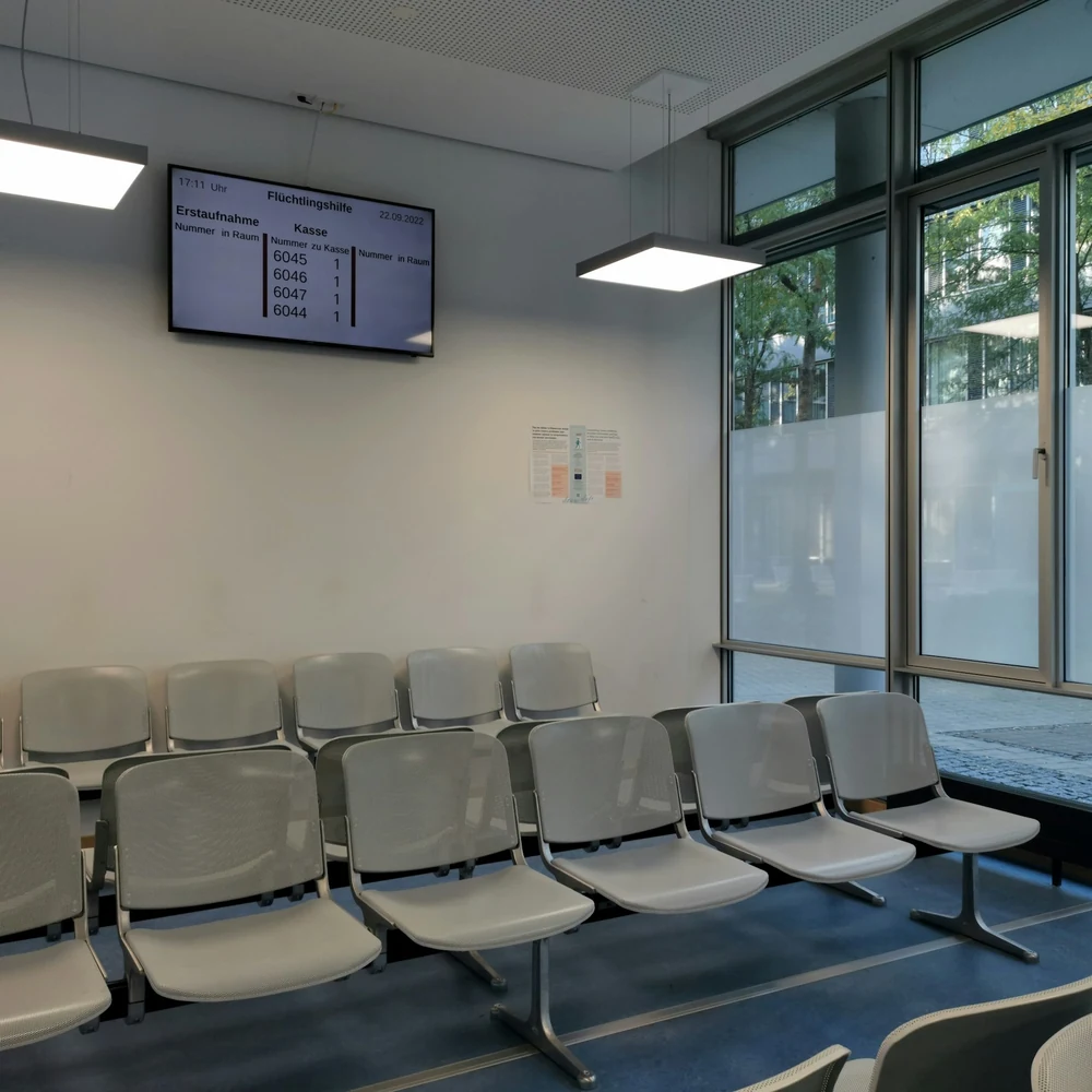 Clinic waiting room with rows of light grey linked chairs, a wall-mounted screen and tall windows