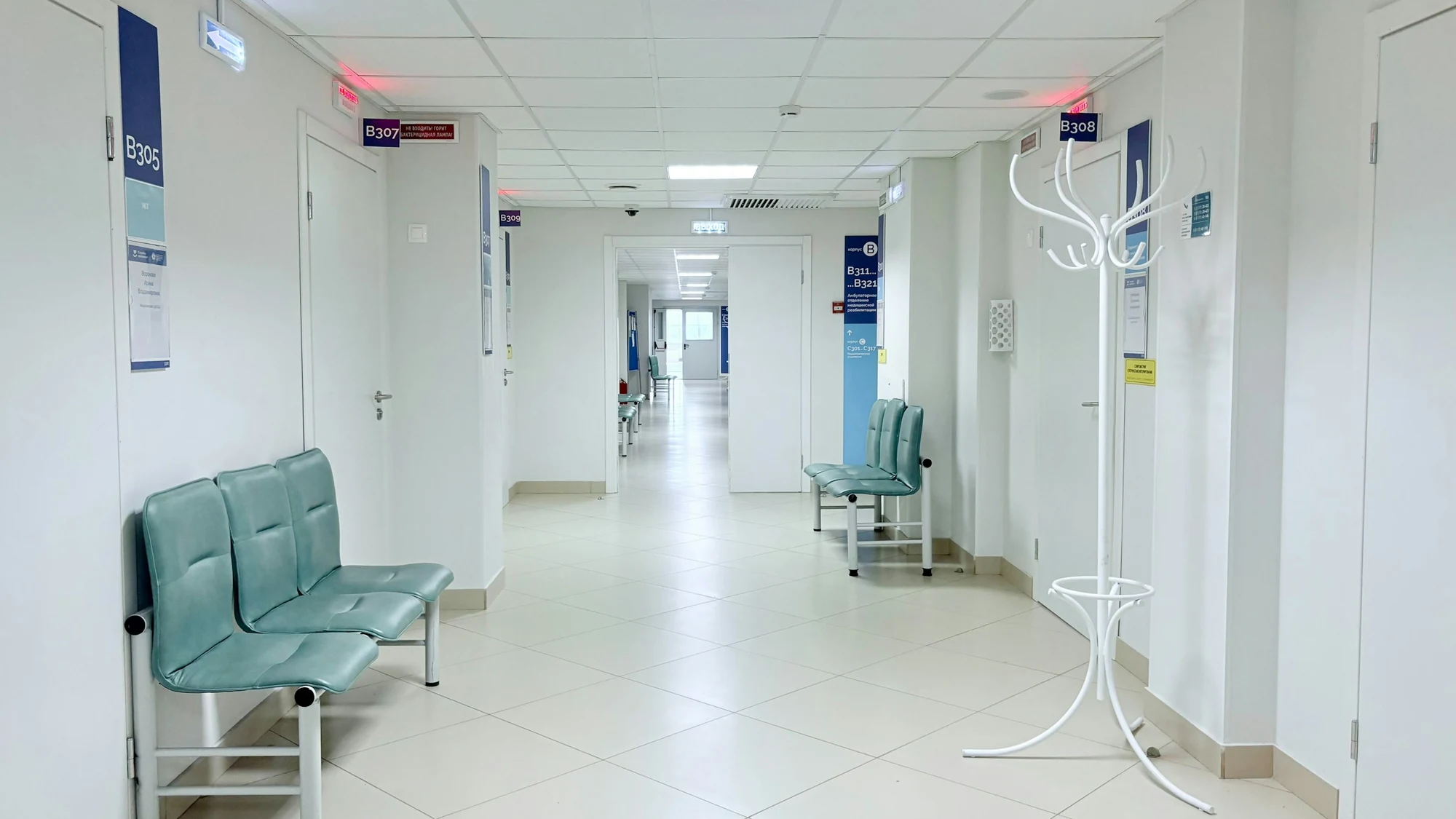 Bright hospital corridor with white tiled walls and floor, doors along both sides and pale blue waiting benches