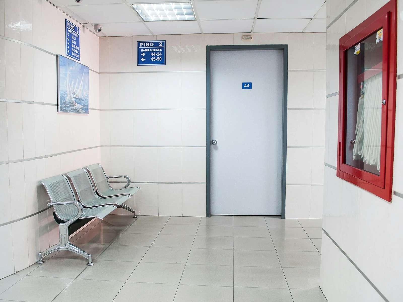 White-tiled hospital waiting corner with a row of metal chairs beside a closed door and a fire hose cabinet