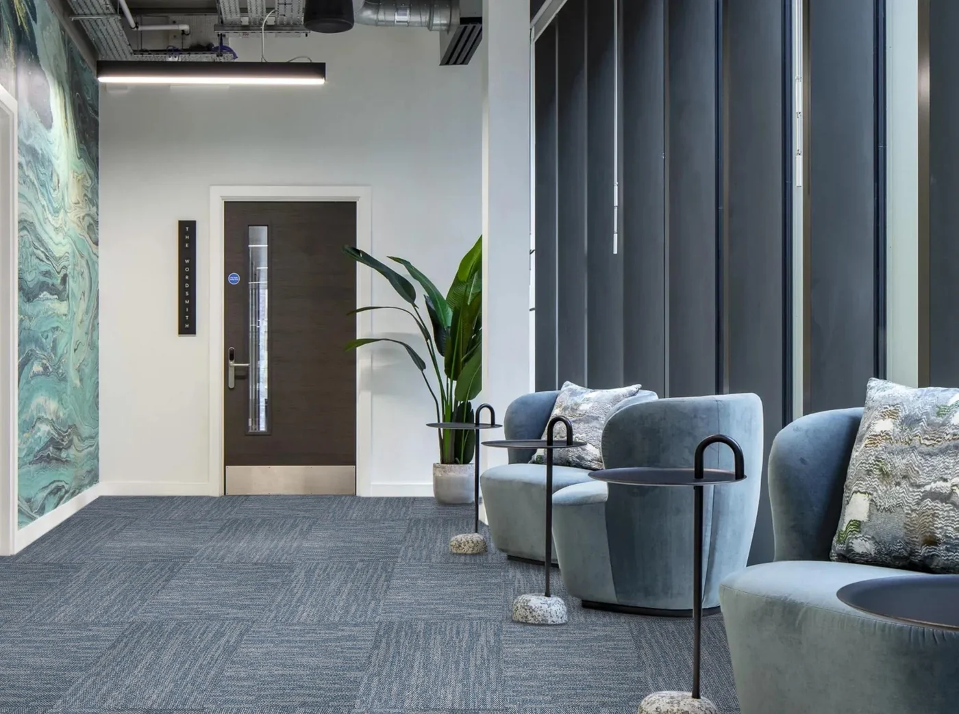 Lift lobby with blue armchairs and a tall plant on grey-blue textured carpet tiles beside a glazed door