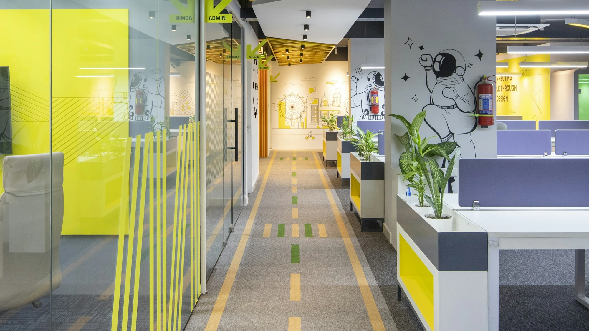 Office corridor carpeted in grey tiles with a yellow and green painted path, glass partitions and a yellow ceiling grid