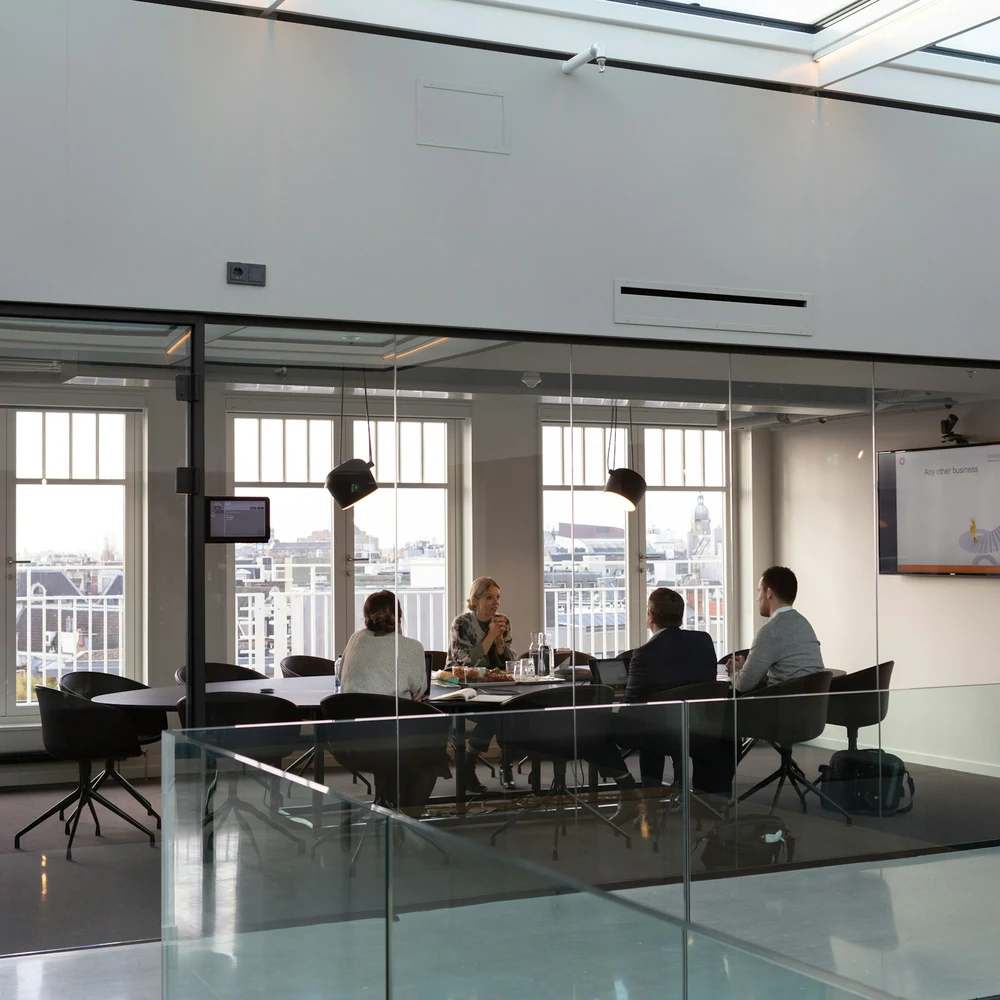 Glass-walled meeting room overlooking an atrium, with people seated at a long table and a screen on the wall