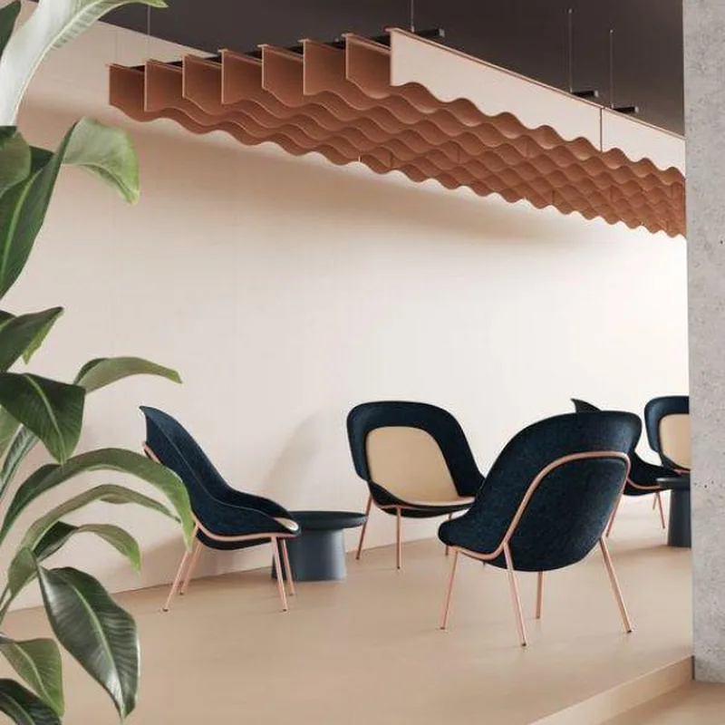 Terracotta wave-shaped acoustic baffle hung from a dark ceiling above navy lounge chairs, with a large leafy plant in front