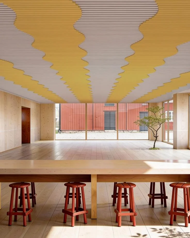 Long timber table with red stools under a ceiling of yellow wave-shaped acoustic baffles, in a bright room with timber floors