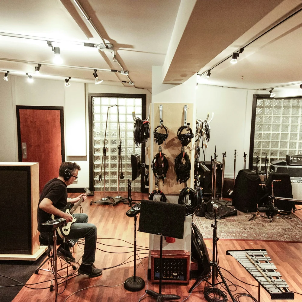 Bright rehearsal room with a guitarist seated among amplifiers, drums and instruments hung on the walls