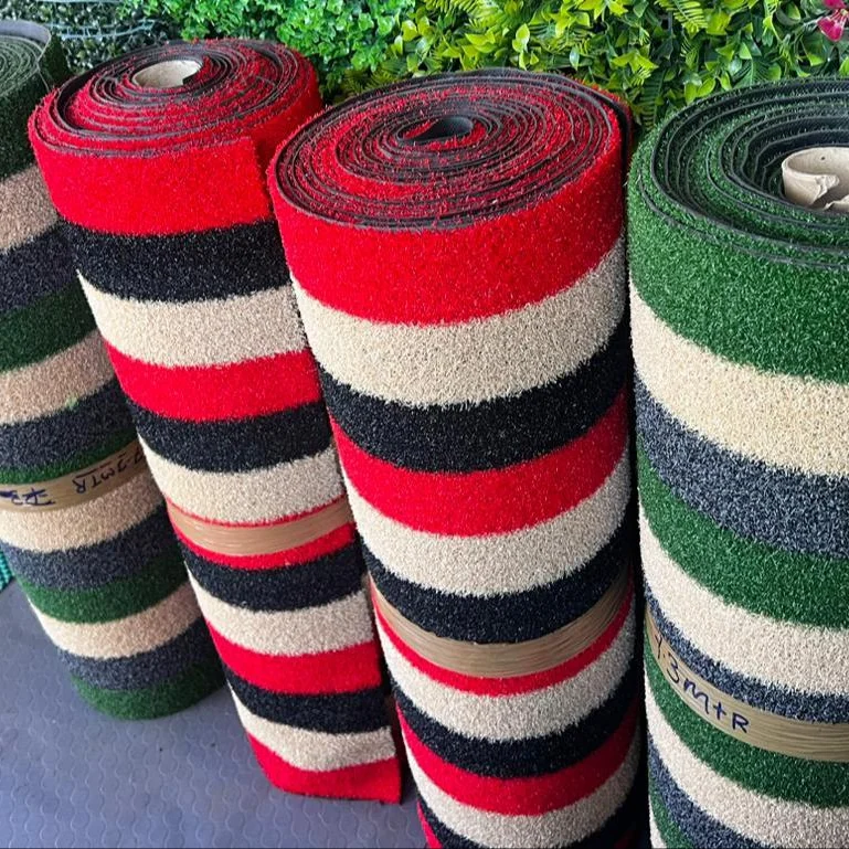 Four rolls of textured grass matting in stripes of green, red, black and cream, standing upright against a hedge