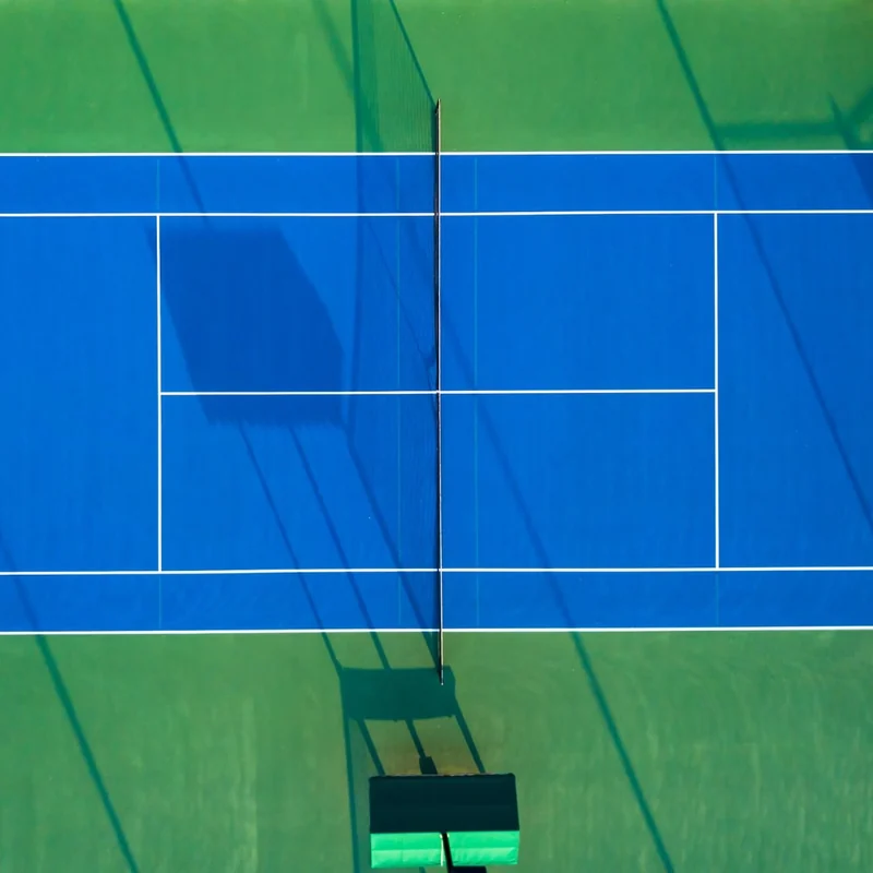 Aerial view of a blue tennis court with white lines and a net, surrounded by green artificial turf